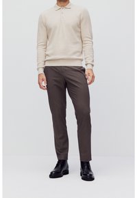 Homme portant un polo beige à manches longues, un pantalon slim marron et des bottes en cuir noires, debout devant un fond clair uni.