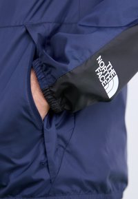 Tumši zila jaka ar melnām akcentiem, ar elastīgiem piedurkņiem un rāvējslēdzēja kabatu. Uz piedurknes drukāts balts logos "The North Face".