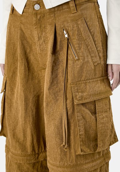 Brązowe, pomarszczone spodnie typu cargo z boczną kieszenią na zamek błyskawiczny, dużą kieszenią z klapą oraz detalem ze sznurkiem, noszone z jasnobeżową bluzką.