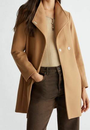 Vrouw draagt een kamelkleurige dubbelgeknoopte jas over een beige geribde trui en donkerbruine jeans, één hand in de zak, neutrale achtergrond.