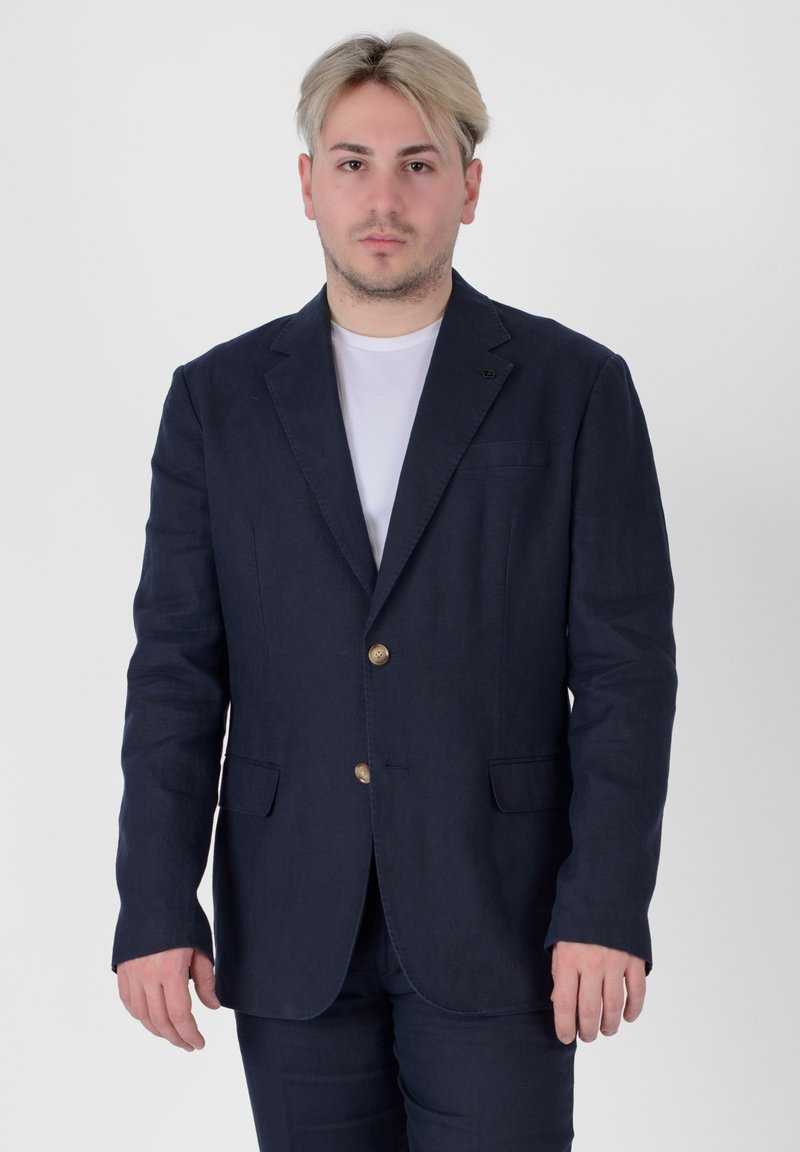 Blazer di lino blu navy con due tasche frontali, due bottoni e rever a punta. Indossato sopra una maglietta bianca, presentato su uno sfondo semplice.