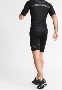 Camiseta y pantalones cortos deportivos negros, con un diseño ajustado, acentos texturizados y rayas horizontales cerca del dobladillo. Llevados con zapatillas negras.