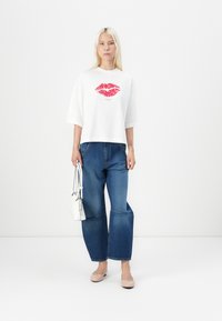White graphic t-shirt with red lip print, paired with blue wide-leg jeans. Model holds a small white structured handbag and wears light flats.