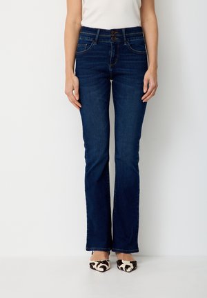 Frau trägt dunkelblaue Bootcut-Jeans mit hoher Taille und doppeltem Knopfverschluss sowie ein weißes Oberteil, steht vor einem einfarbigen weißen Hintergrund.