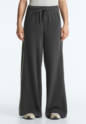 Personne portant un pantalon de survêtement gris foncé à jambes larges avec une taille élastique et un cordon de serrage, associé à des baskets de couleur claire.