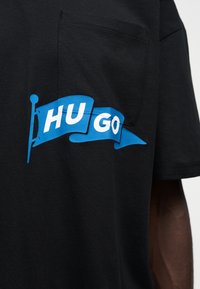 Μαύρο μπλουζάκι με τσέπη στο στήθος και μπλε σημαία με λευκό κείμενο "HUGO" στο μπροστινό μέρος.