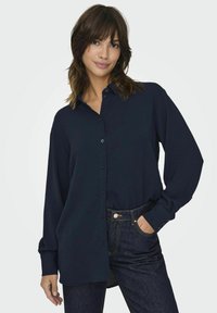 Navy blue button-up shirt with long sleeves, textured fabric, and a relaxed fit, paired with dark denim jeans and a visible button detail.