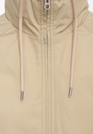 Beige jas met een hoge kraag, ritsluiting en trekkoorden. Heeft een soepele, matte textuur en accenten van zilverkleurig hardware.
