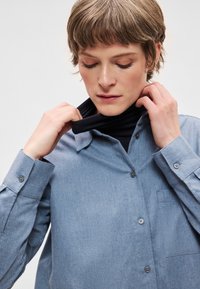 Světlé modré košile z tkané látky s náprsní kapsou a dlouhými rukávy s manžetami na knoflíky, nošené přes černý rolák.