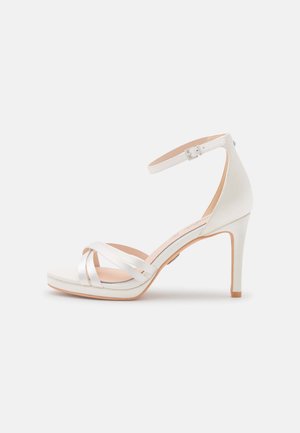 White high-heeled sandal with an ankle strap, soft leather upper, and crossed strap design. Features a light brown outsole.