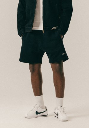 Mann trägt schwarze Shorts, weiße Sportsocken, weiße Nike-Sneaker, schwarze Jacke und weißes T-Shirt, steht mit den Händen in den Taschen.