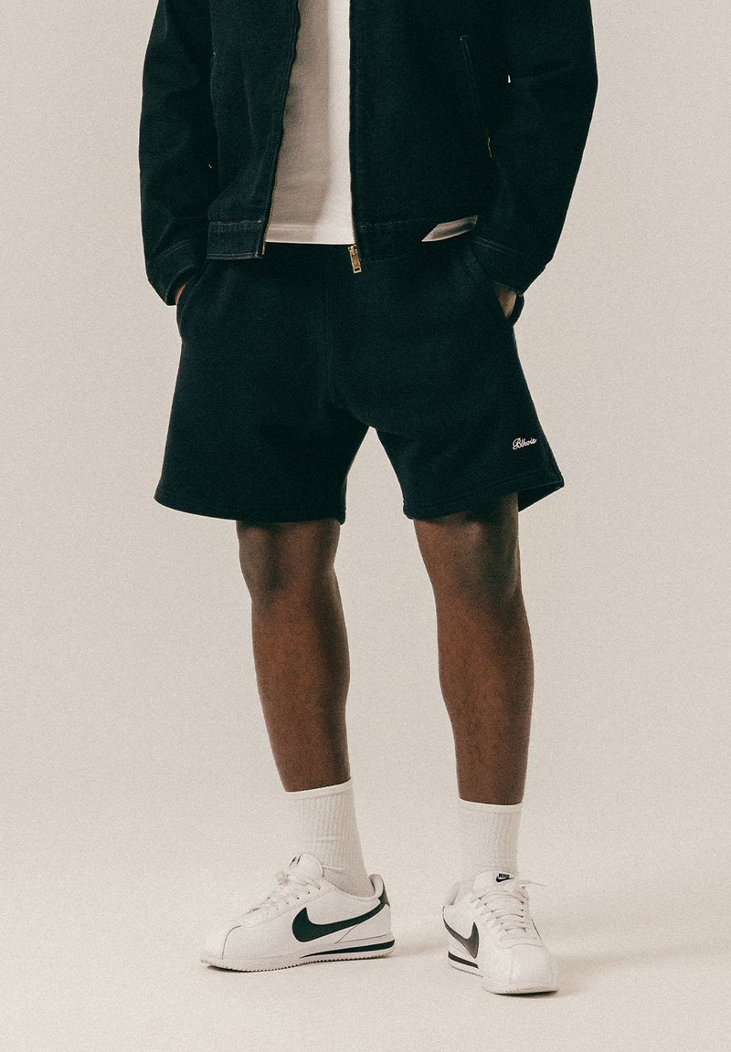 Homme portant un short noir, des chaussettes blanches montantes, des baskets blanches Nike, une veste noire et un T-shirt blanc, debout les mains dans les poches.