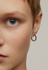 Pendiente de aro metálico en tonos oscuros con detalle texturizado y un pequeño acento brillante. Usado en la oreja izquierda, vista en primer plano de la cara del modelo.