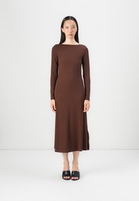 Brun ribbestrikket kjole med langt erme, båthals og en kroppsnær silhuett; kombinert med enkle svarte åpen-tå sandaler.