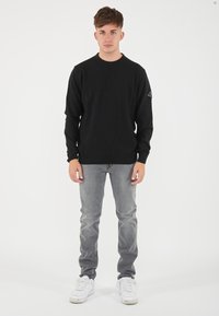 Maglione nero a maniche lunghe con colletto rotondo e finitura testurizzata, abbinato a jeans grigi chiari e sneakers bianche.