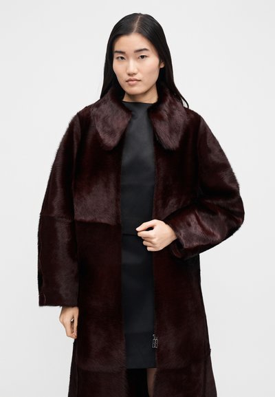 Moteris apsirengusi tamsiai bordo dirbtinio kailio paltu ant juodos suknelės, stovinti priešais paprastą šviesiai pilką foną.