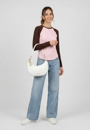 Mladá žena stojící, oblečená v růžovo-hnědém raglánovém tričku, světle modrých širokých džínách a bílých teniskách, držící bílou kabelku s pleteným popruhem.