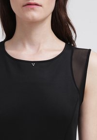 Kobieta w bezrękawowej czarnej bluzce z przezroczystą siateczkową wstawką na ramieniu oraz małym trójkątnym metalowym zdobieniem u dekoltu.