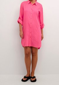 Jasnoróżowa lniana sukienka koszulowa z zapięciem na guziki z przodu, długimi rękawami podwiniętymi oraz zaokrąglonym dołem. Zestawiona z czarnymi płaskimi sandałami.