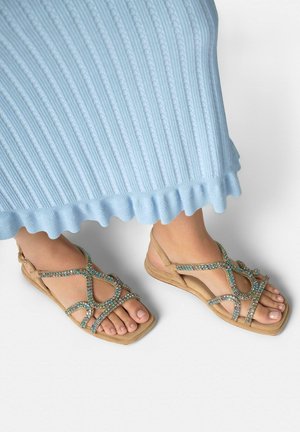 Pies con sandalias planas beige con tiras multicolores brillantes, parcialmente cubiertos por una falda azul claro acanalada con un dobladillo con volantes.