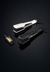 Deux fers à lisser, l'un blanc et l'autre noir, avec des designs élégants et des bords arrondis, accompagnés d'une petite bouteille noire avec des accents dorés.