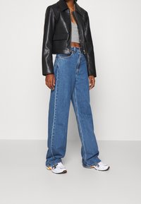 Veste en cuir noire raccourcie, débardeur gris ajusté, jean en denim bleu à jambes larges, et baskets blanches avec des accents orange, sur un fond neutre.