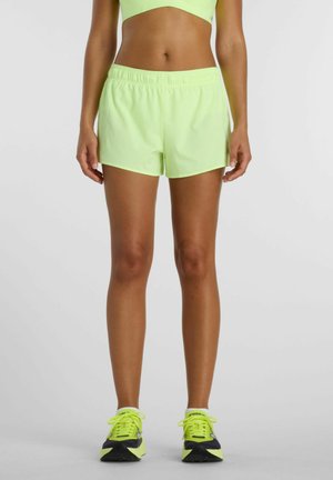 Femme portant un short de sport vert fluo, un crop top assorti et des chaussures de course vert fluo et noires, debout devant un fond uni.