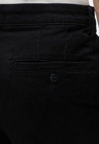 Gros plan de la poche arrière d'un pantalon noir avec fermeture par bouton et détails de couture visibles.