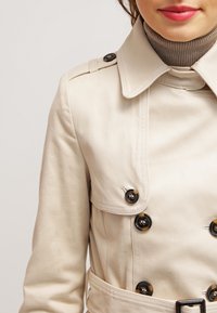 Trench-coat beige avec un col structuré, une fermeture à double boutonnage et une taille ceinturée. Texture lisse et détails de couture prononcés.