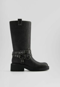 Bota alta hasta la rodilla de cuero sintético negro con tacón grueso, doble correa con hebilla y detalles de ojales metálicos. Textura lisa y puntera redondeada.