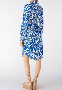Abito con motivo floreale in blu e bianco, dotato di maniche lunghe, una cintura in vita e un orlo curvato. Indossato con sandali con cinturini color sabbia.