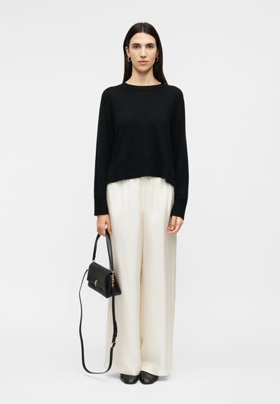 Woman with long dark hair wearing a black sweater and white wide-leg pants, holding a black handbag with a shoulder strap.