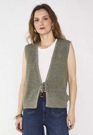 Donna con gilet grigio a maglia senza maniche annodato in vita sopra una camicia bianca, jeans blu, orecchini dorati e braccialetti, in piedi contro uno sfondo bianco.