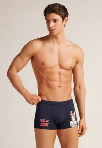 Bóxers de color azul marino hechos de algodón, con un diseño impreso de Tom y Jerry en el lado izquierdo y texto rojo. Forma ajustada con cinturilla elástica.