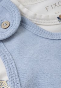 Bodysuit en coton bleu clair avec une texture douce, doté d'un col arrondi et d'une fermeture à bouton. Tissu blanc en dessous avec des accents de design subtils.