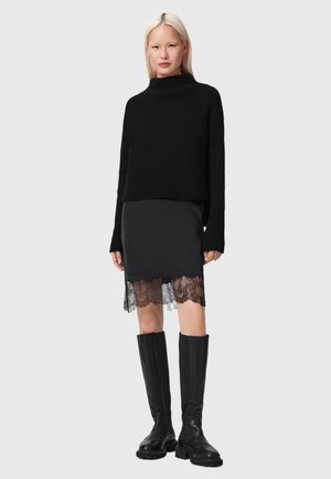 Vrouw draagt een zwarte geribbelde coltrui, zwarte rok met kanten zoom en kniehoge zwarte leren laarzen, staand tegen een effen achtergrond.