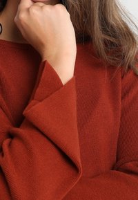 Sweter w kolorze rdzy z dzianiny, z rozszerzającymi się rękawami i prążkowaną fakturą, podkreślający miękkość i ciepło materiału.