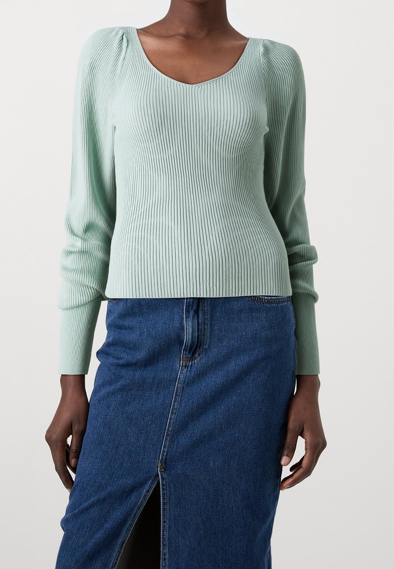 Femme portant un haut côtelé à manches longues vert clair et une jupe en denim bleu avec une fente à l'avant, debout devant un fond uni.