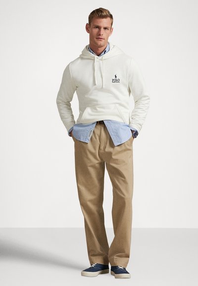 Κρεμ φούτερ με κουκούλα και λογότυπο, ανοιχτό μπλε πουκάμισο από κάτω, μπεζ παντελόνι και μπλε αθλητικά παπούτσια. Καθημερινό στρωμένο σύνολο με λεπτομέρεια τσέπης στο φούτερ.