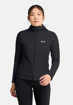 Vrouw die een zwarte Jack Wolfskin jas met rits en capuchon draagt, staand tegen een effen lichte achtergrond, licht naar rechts kijkend.