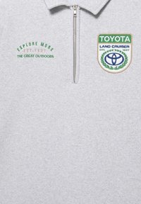Polo grigio con zip e testo ricamato "EXPLORE MORE" e "THE GREAT OUTDOORS". Presenta una toppa con il design di una corona d'alloro e la scritta Toyota Land Cruiser.