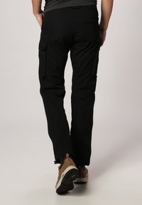 Un pantalon cargo noir présente une coupe slim avec des poches et un léger stretch. La taille est élastique et ils sont associés à des chaussures marron.