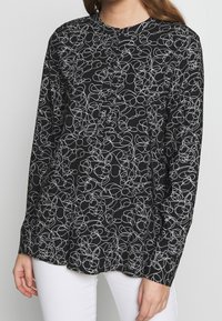 Chemise noire à manches longues avec un motif de lignes abstraites blanches, col rond et fermeture par boutons. Tissu doux, coupe décontractée.