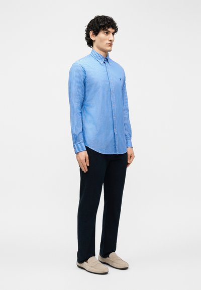 Άνδρας που στέκεται, φοράει μπλε ριγέ πουκάμισο με κουμπιά, μαύρο παντελόνι και μπεζ μοκασίνια, σε απλό φωτεινό φόντο.