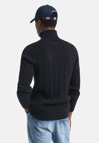 Schwarzer Kabelstrickpullover mit hohem Kragen und gerippten Bündchen, kombiniert mit hellblauen Jeans. Das Outfit hat einen lässigen Schnitt und ein strukturiertes Design.