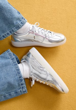 Personne portant des chaussettes blanches, un jean bleu et des baskets argentées métalliques assise sur un sol texturé jaune moutarde.
