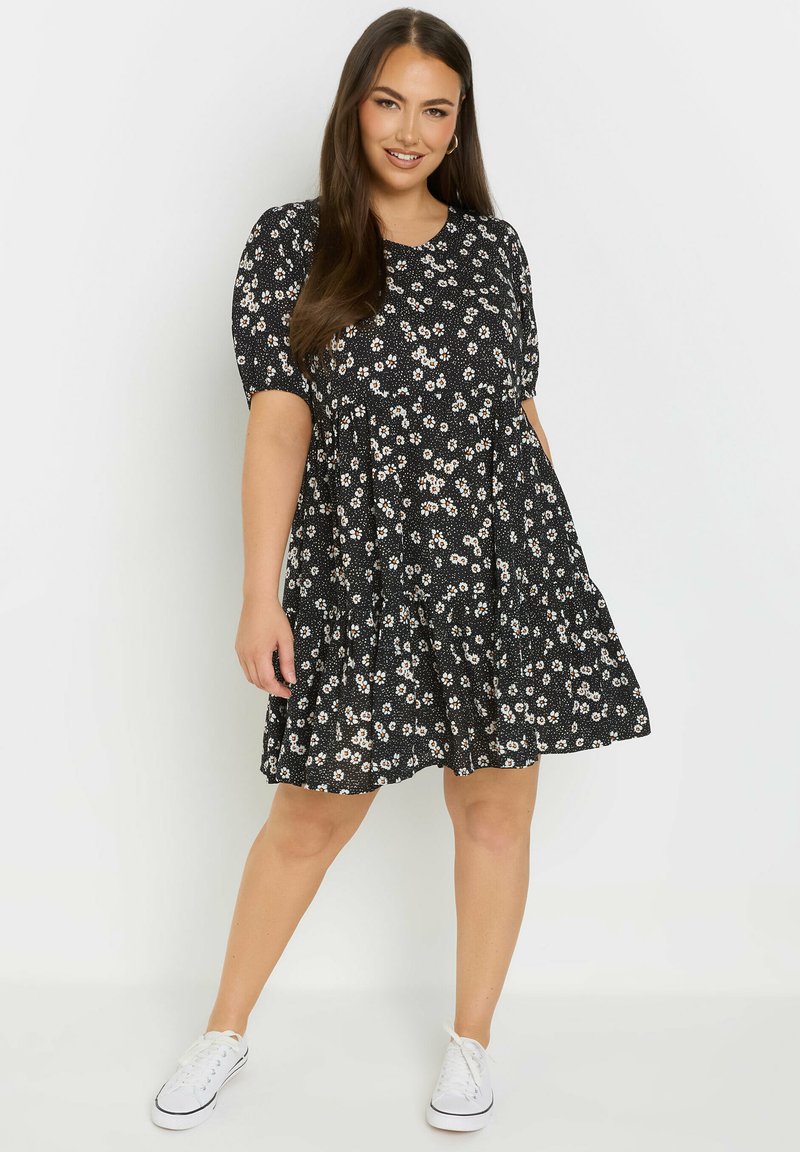 Mekko, jossa on musta kukkakuosi, lyhyet hihat ja kerroksellinen hame, jossa on pieniä valkoisia ja vaaleanpunaisia kukkia. Yhdistetty valkoisiin lenkkareihin.