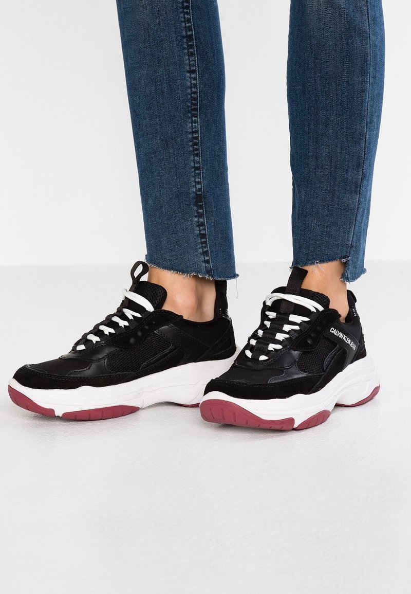 Des chaussures de sport noires avec des accents blancs, composées d'un mélange de filet et de daim, avec une épaisse semelle blanche ornée de détails rouges et des lacets noirs.