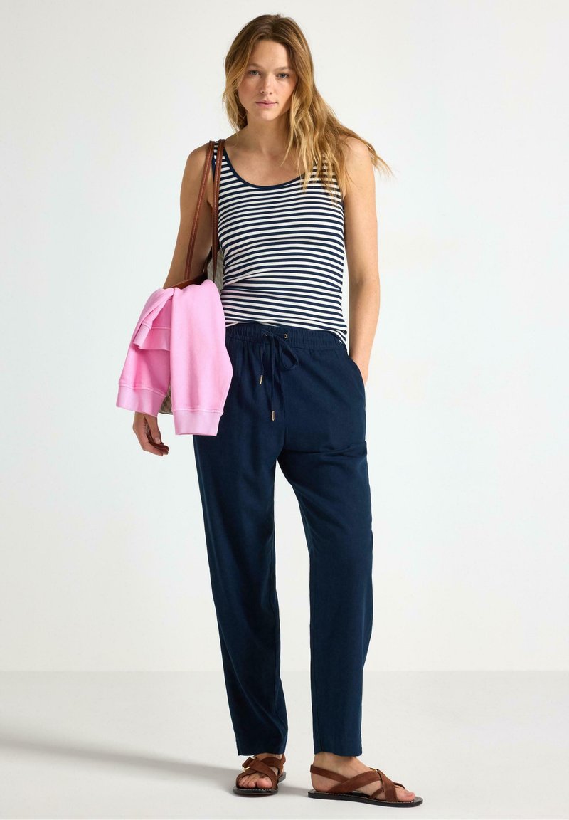 Canotta a righe blu navy e bianche, pantaloni blu navy con cordoncino, felpa rosa chiaro appoggiata sul braccio e sandali marroni. Outfit semplice e informale.