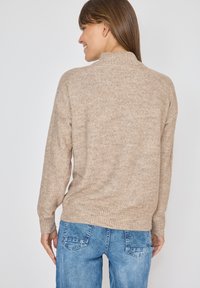 Beżowy sweter dzianinowy z wysokim kołnierzem, ściągaczami przy mankietach i u dołu. Miękka faktura, lekko luźny krój, zestawiony z jasnoniebieskimi dżinsami.